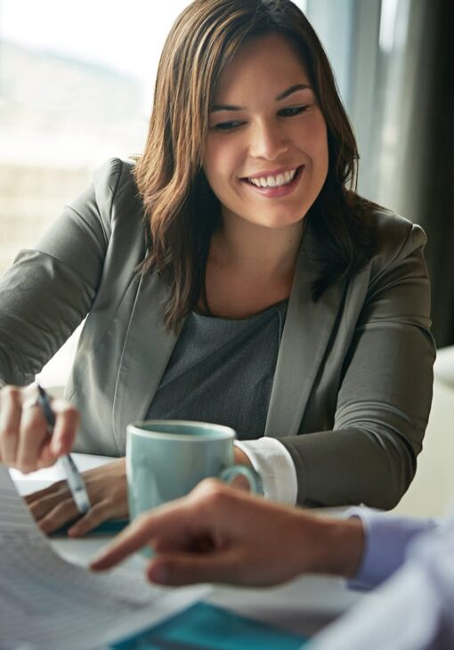 Smiling woman in a blazer holding a pen and coffee mug, reviewing documents with a colleague in a well-lit office.