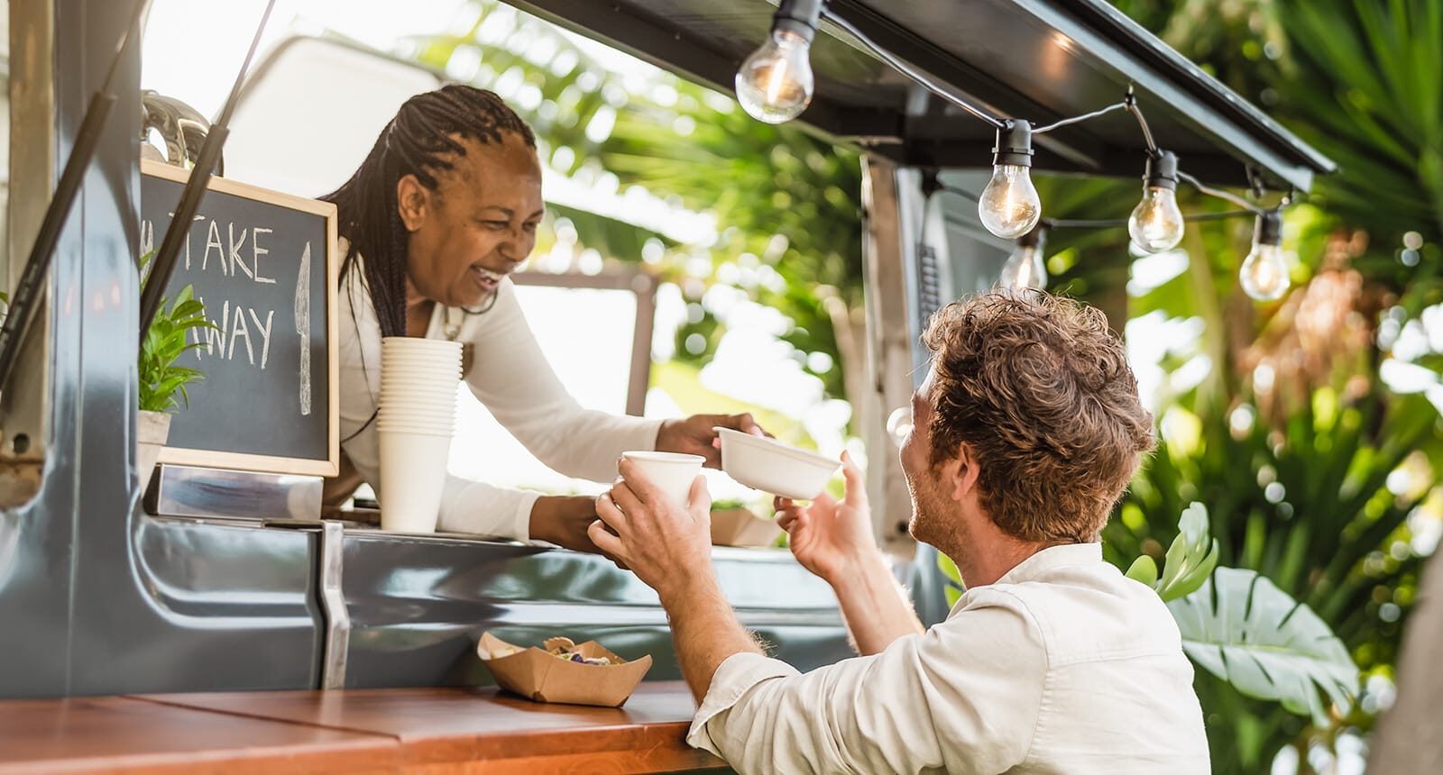 Smiling woman serving food and drinks from a food truck to a male customer under hanging lights, surrounded by lush greenery.