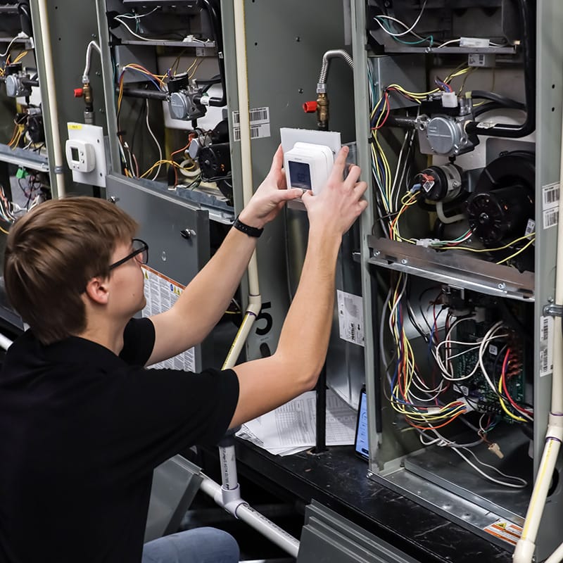 Student in safety glasses installs a wall-mounted thermostat while working on open HVAC units with exposed wiring in a classroom lab.