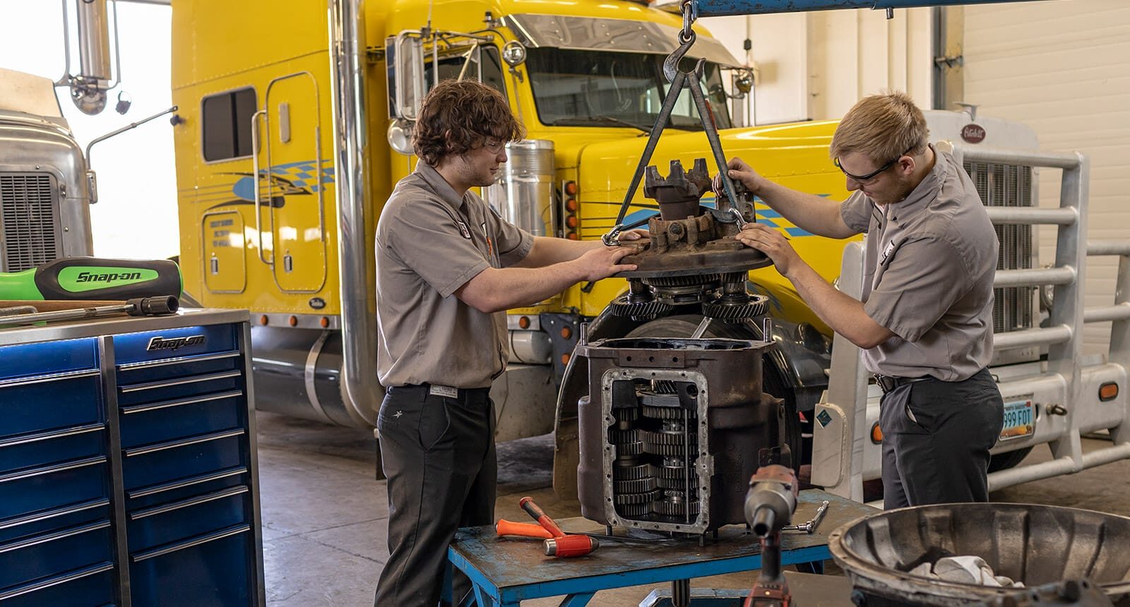 Two diesel students in a truck bay working together to hoist a large gear assembly into place, with semi-trucks and tools in the background.