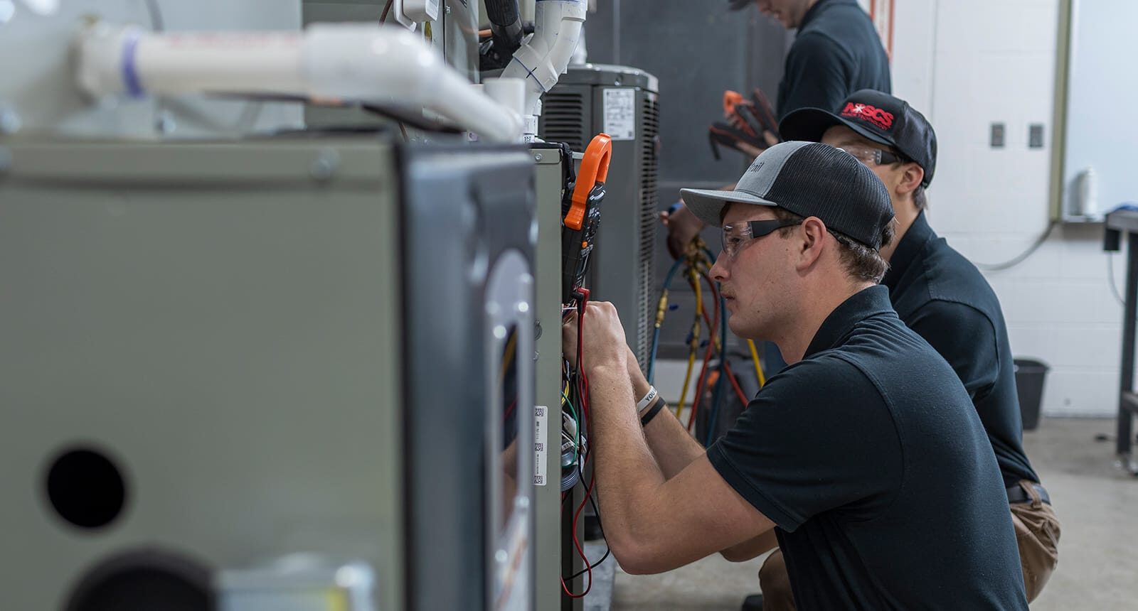 Students wearing black shirts and safety glasses work on HVAC units, using multimeters and examining wiring in a hands-on technical lab environment.