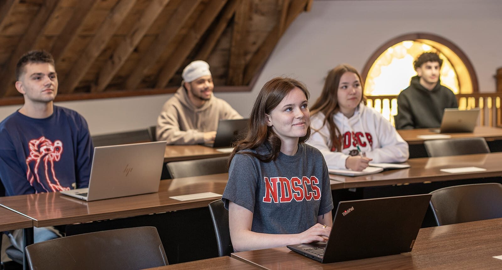 Students seated at desks with laptops in a classroom featuring wood-paneled ceilings and a stained glass window in the background.