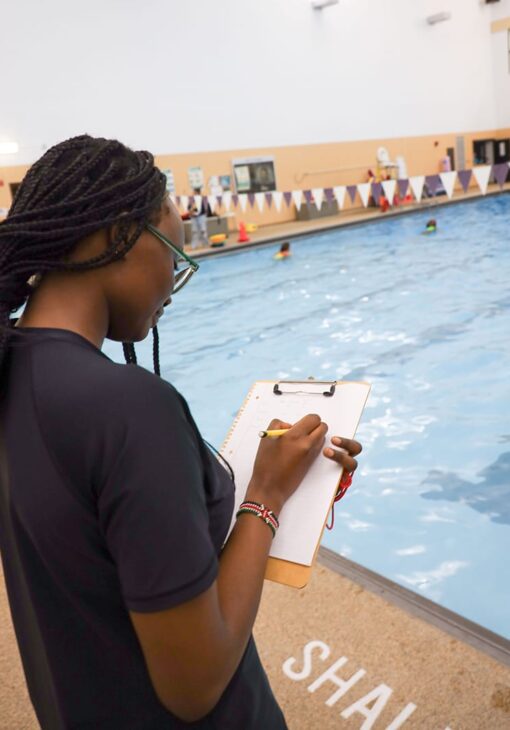 Woman with braided hair writing on a clipboard beside an indoor swimming pool, observing swimmers during a class or training session.