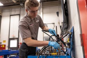 Student in safety glasses and gloves working with hydraulic hoses on a training station in a workshop.