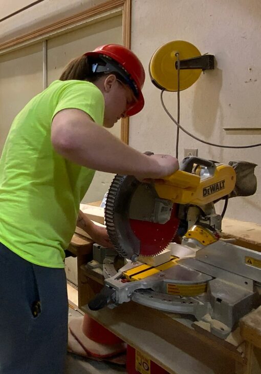 Person in a red hard hat and neon yellow shirt operating a DeWalt miter saw in a woodshop setting.