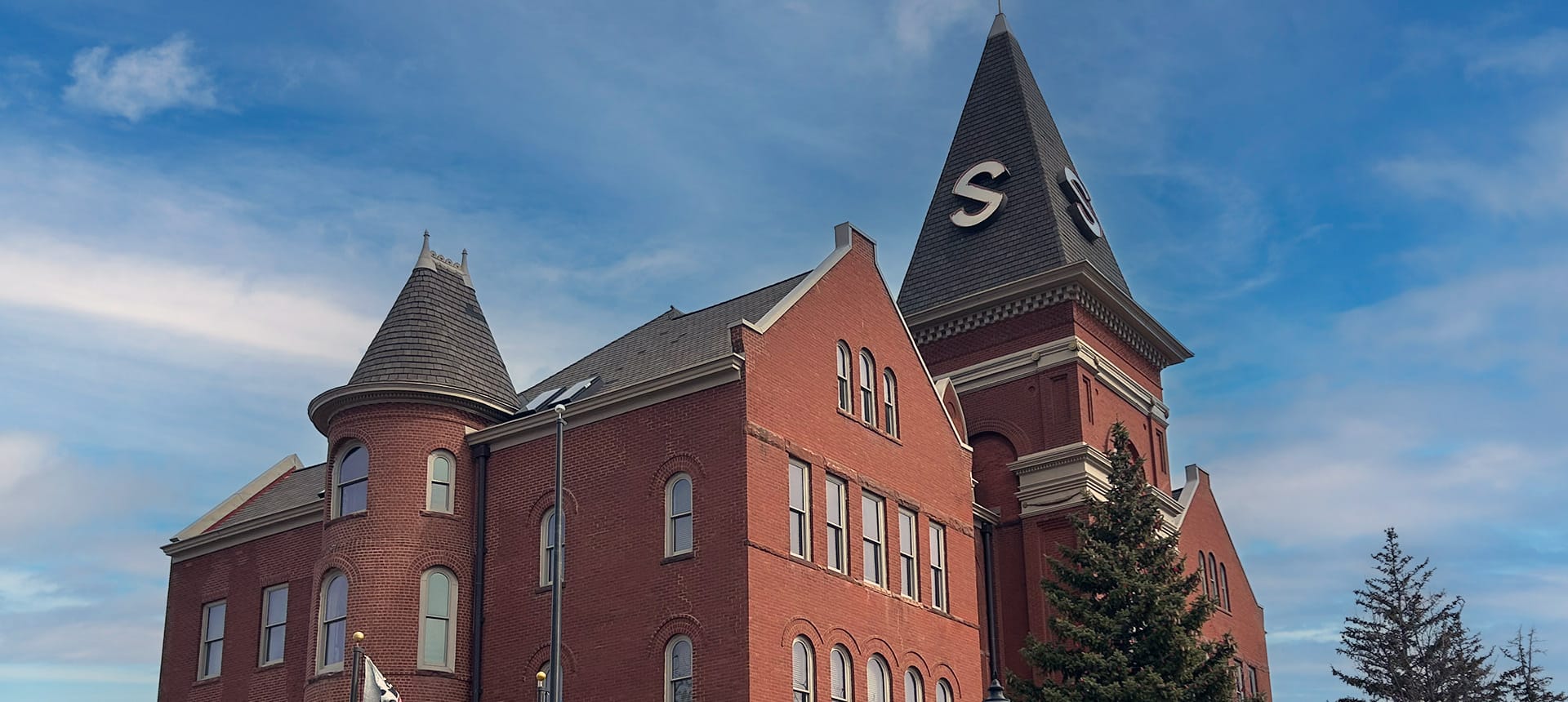 Historic red brick campus building with two towers, arched windows, and a steep roofline under a blue sky.