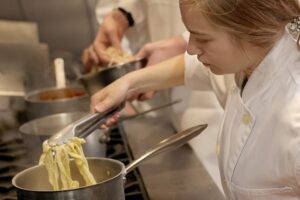Student in a white chef’s coat using tongs to transfer cooked pasta from a pot on the stove, with other pots and a second student visible in the background.