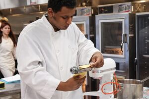 Student in a white chef’s coat using a pasta roller attachment on a stand mixer to flatten fresh pasta dough in a commercial kitchen.