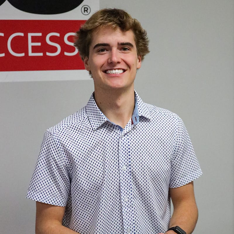 Smiling young man in a short-sleeved, patterned button-up shirt standing in front of a wall with partial red and black signage.