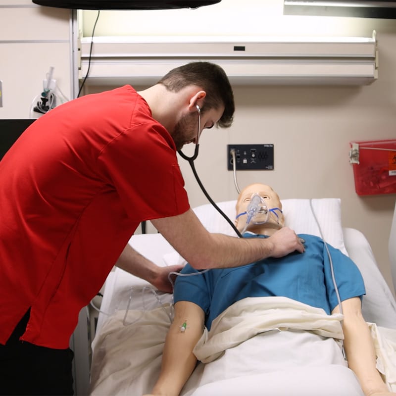 Nursing student in red scrubs using a stethoscope to listen to the chest of a medical mannequin wearing an oxygen mask.