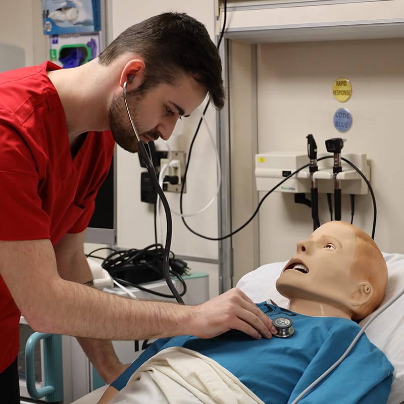Male nursing student in red scrubs using a stethoscope to assess a child-sized medical mannequin in a simulation lab.
