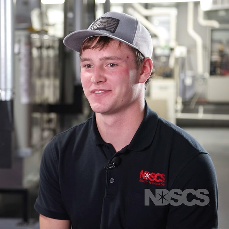 Smiling student in an NDSCS HVAC/R Technology polo and baseball cap sits for an interview in a lab setting.