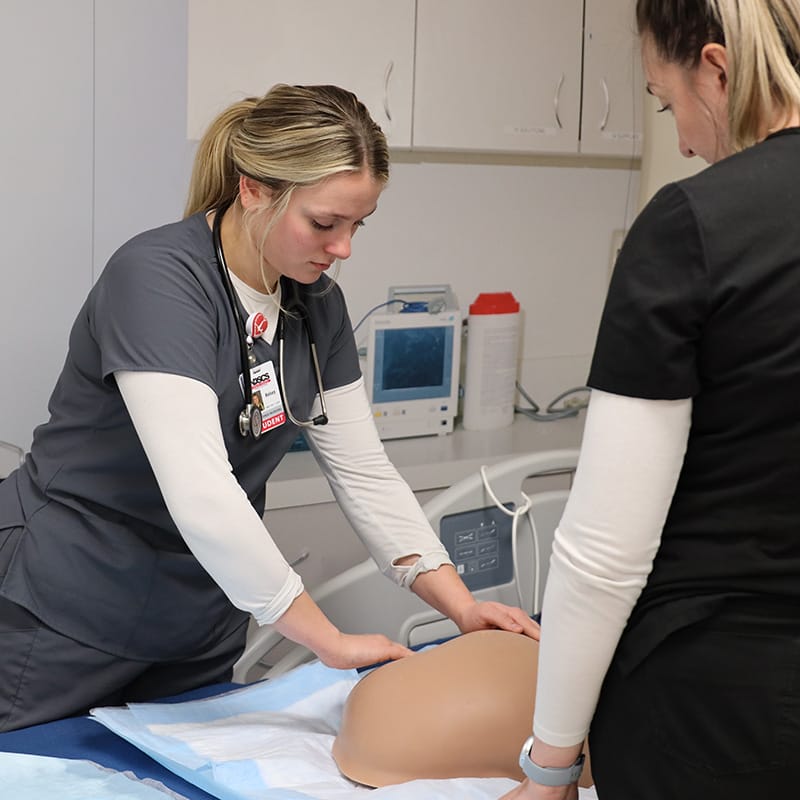 Nursing student in gray scrubs practicing abdominal assessment on a medical mannequin while instructor observes.