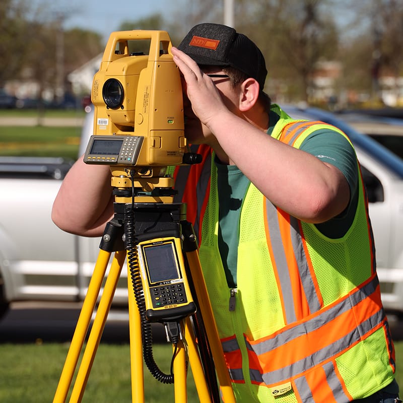 Student in a safety vest and hat uses a total station surveying instrument on a tripod outdoors.