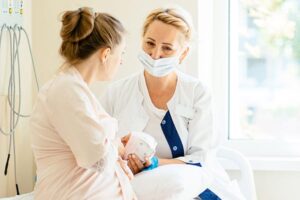 Nurse in a mask offering support to a new mother holding her newborn in a hospital room with bright window light.