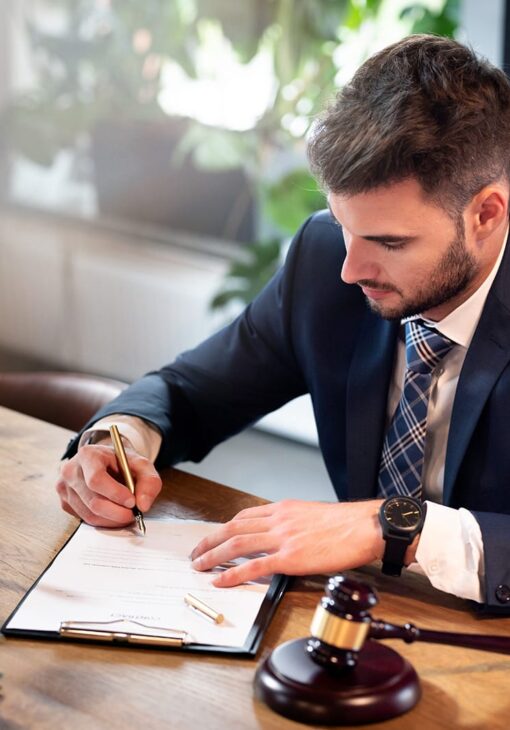 Man in a suit signing a document at a desk with a gavel, representing a legal or law-related setting.