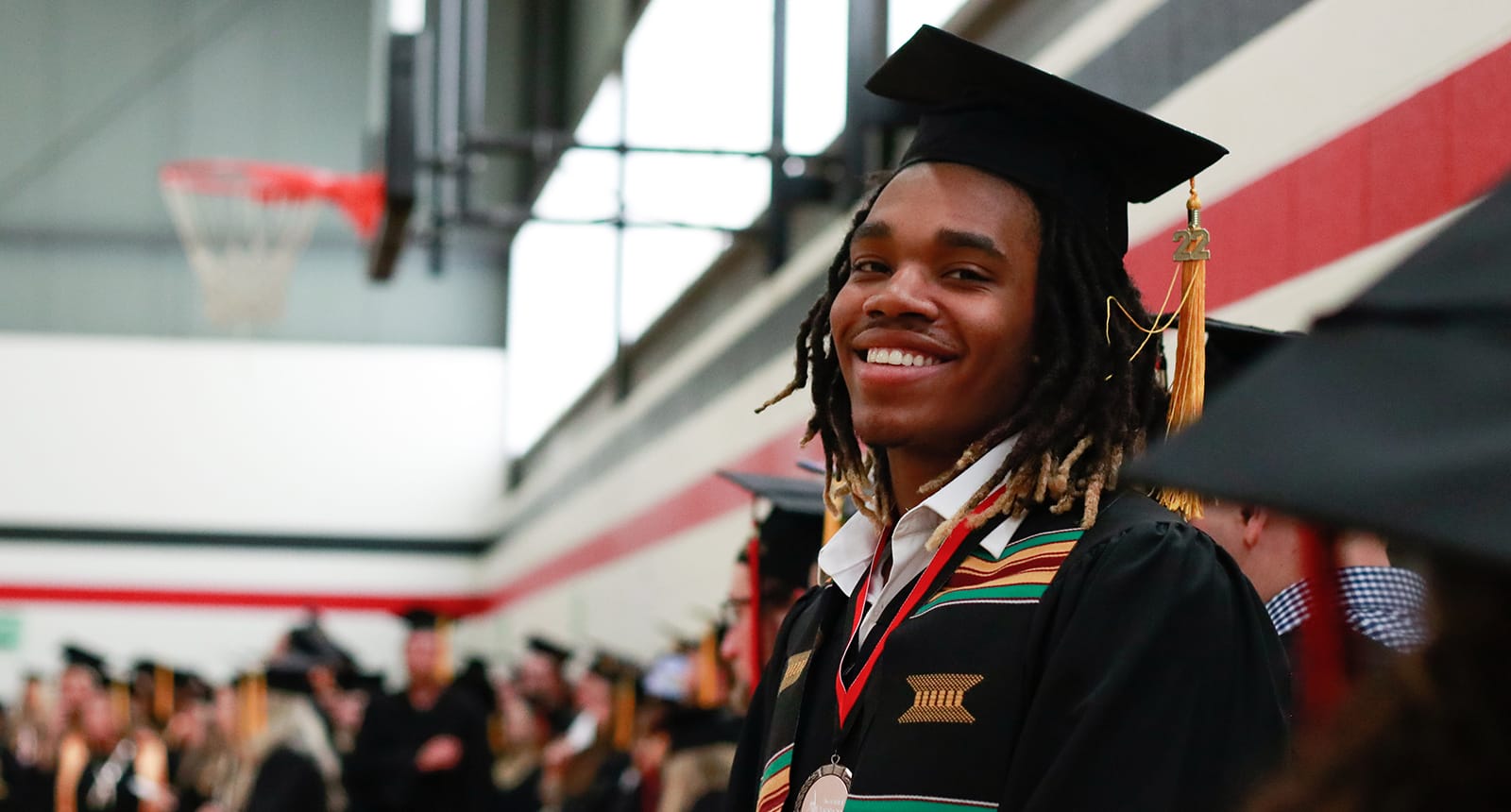 Smiling graduate in cap and gown standing in a gymnasium during a commencement ceremony