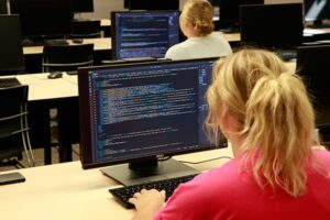 Student with blonde hair in a ponytail working at a desktop computer displaying code in a classroom filled with computers and other students.