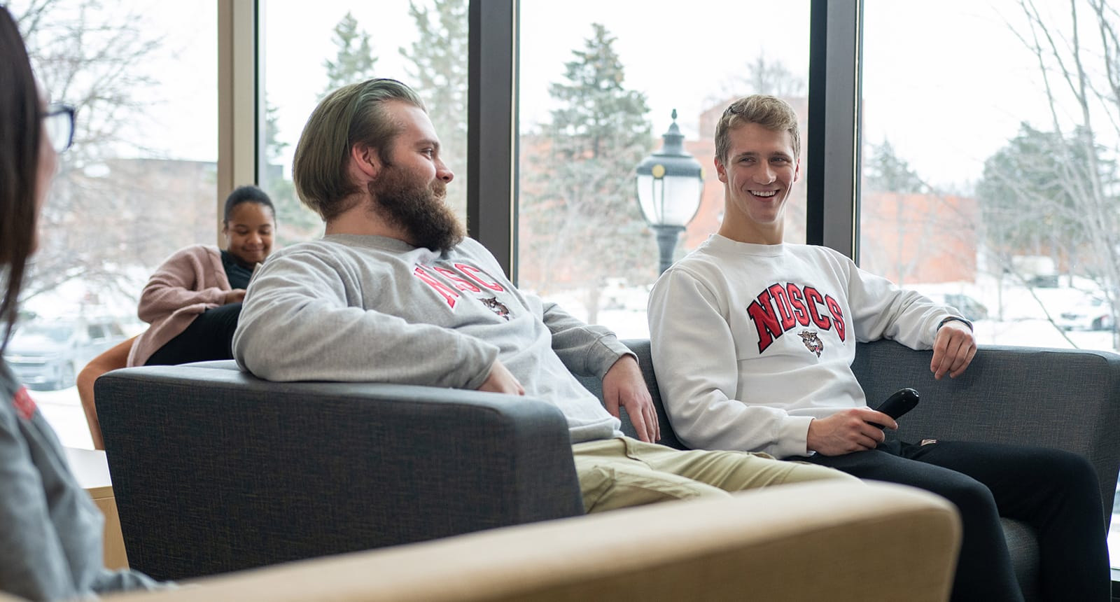 Two smiling students wearing NDSCS sweatshirts sit on a couch inside a bright campus lounge with snowy trees visible through the windows