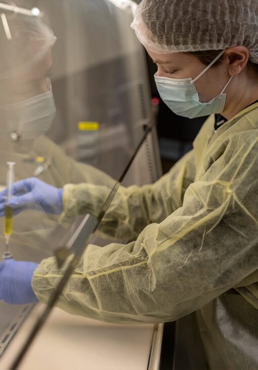 Student in protective gear and mask working inside a sterile pharmacy lab hood, drawing yellow liquid into a syringe from a vial.