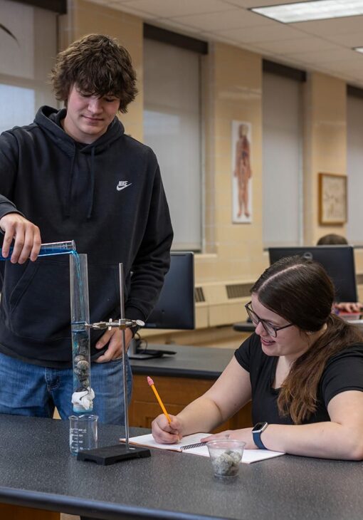 Two students working together on a science experiment, with one pouring liquid into a tall cylinder containing rocks while the other takes notes at a lab table.