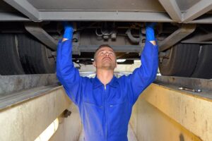 Mechanic in blue coveralls and gloves inspecting the underside of a vehicle from a service pit.