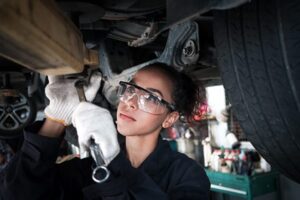Focused female automotive technician in safety goggles and gloves working underneath a vehicle with a wrench.