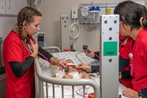 Nursing students in red scrubs using stethoscopes on a training infant mannequin in a hospital crib during a pediatric simulation exercise.