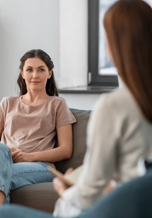 Young woman sitting on a couch with a calm expression, speaking to another woman holding a notebook in a counseling session.