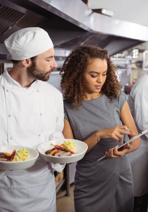 Chef holding two plated meals while a woman in a dress points at a clipboard in a commercial kitchen.