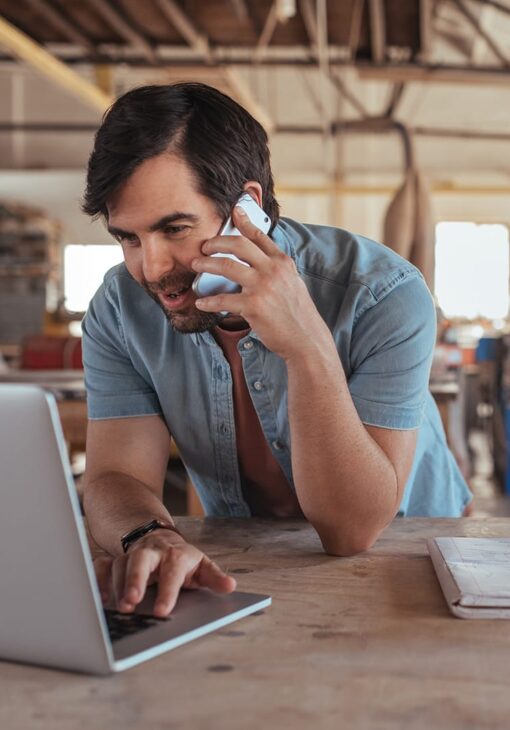 Man in a casual button-up shirt talking on a smartphone while working on a laptop in a workshop or studio setting.