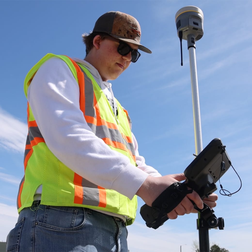 Student wearing a safety vest and cap uses a handheld GPS surveying device outdoors under a clear blue sky.