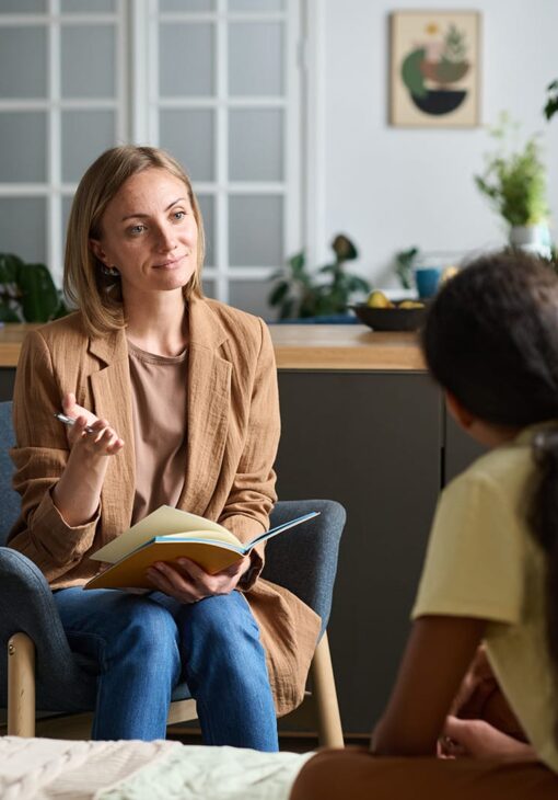 Woman holding a notebook and pen while speaking with another person in a casual indoor setting with plants and modern decor.