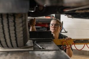 Student wearing safety glasses working underneath a vehicle on a lift, focusing on the steering components.