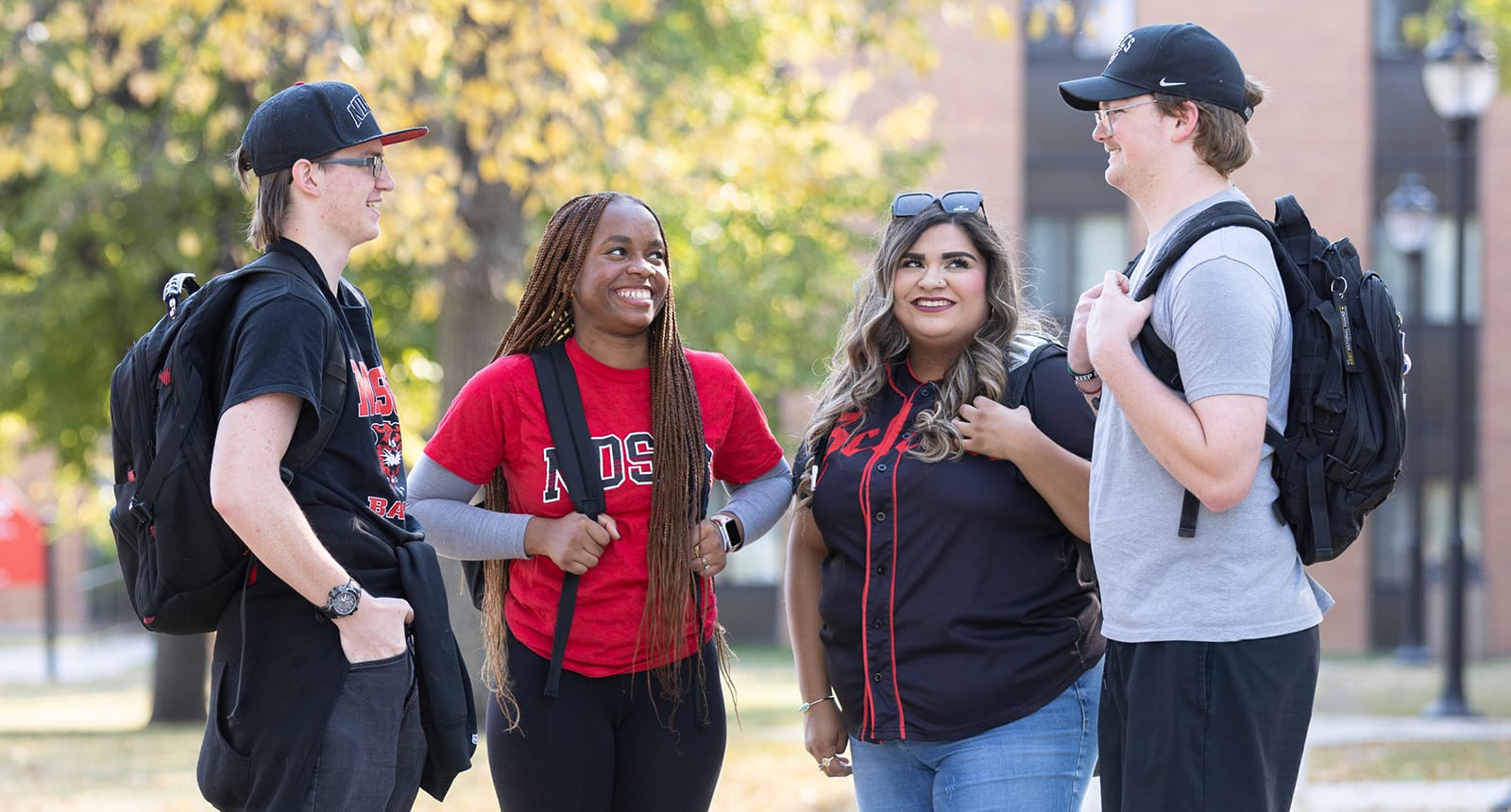 Four students with backpacks stand outside on campus smiling and chatting on a sunny day, with trees and a brick building in the background.