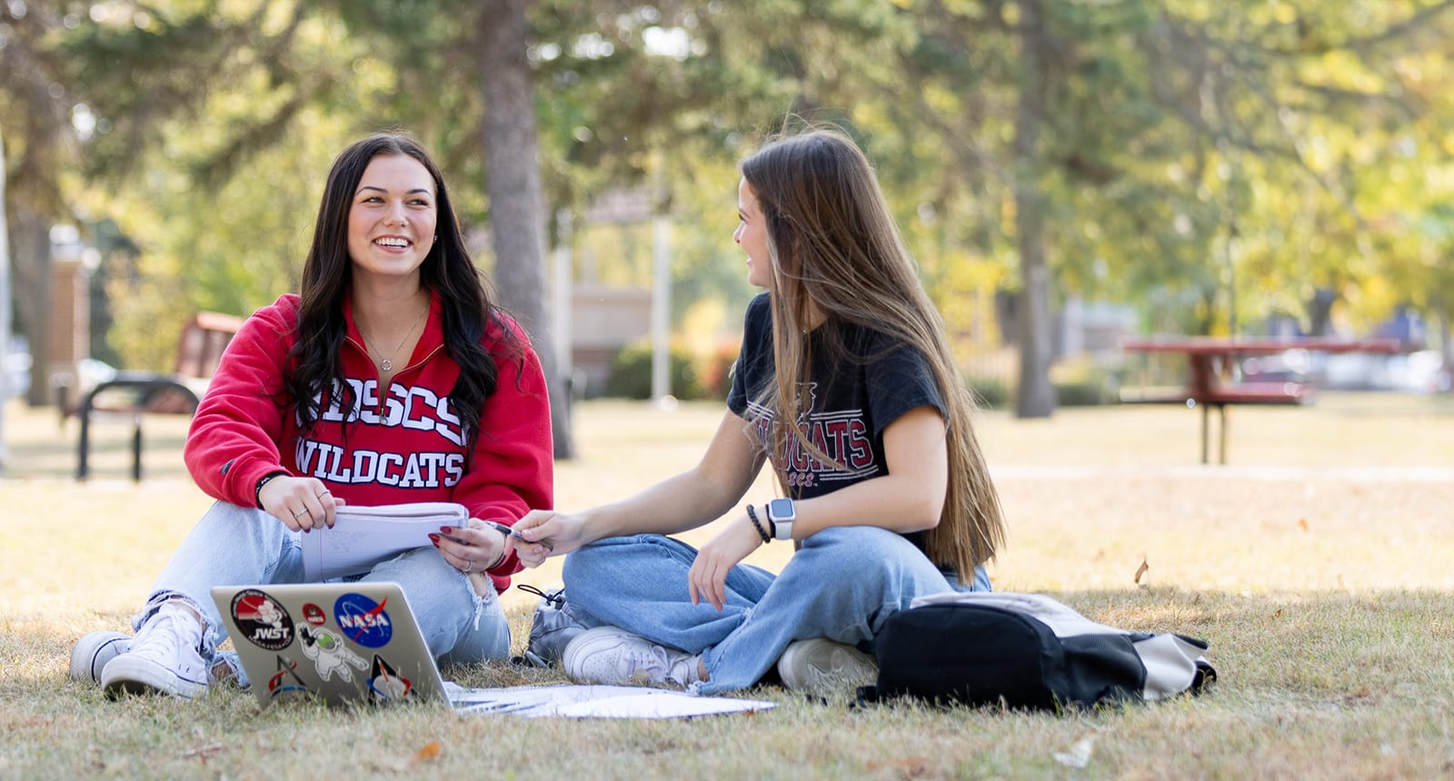 Two students sit cross-legged on the grass, smiling and studying together on a sunny day, with laptops, notebooks, and backpacks in front of them.