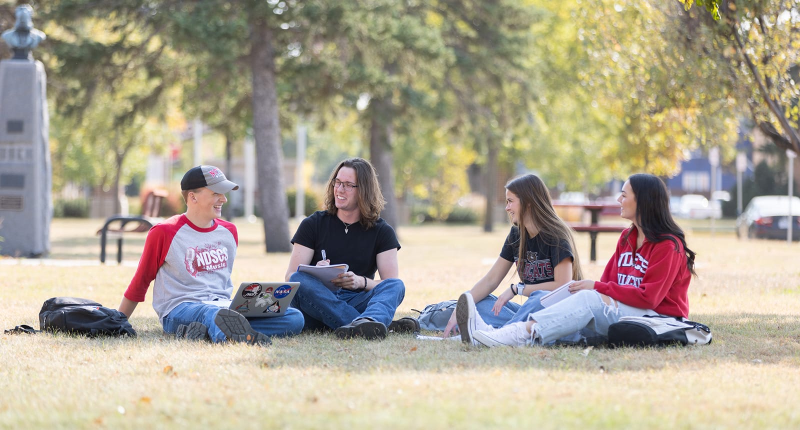 Group of four college students sitting in a circle on the grass, laughing and studying together on a sunny campus lawn
