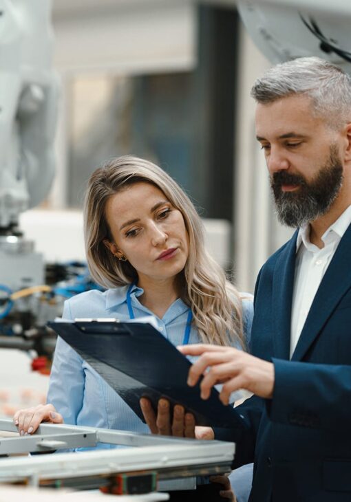 Man and woman reviewing information on a clipboard while standing next to industrial machinery in a manufacturing or tech facility.