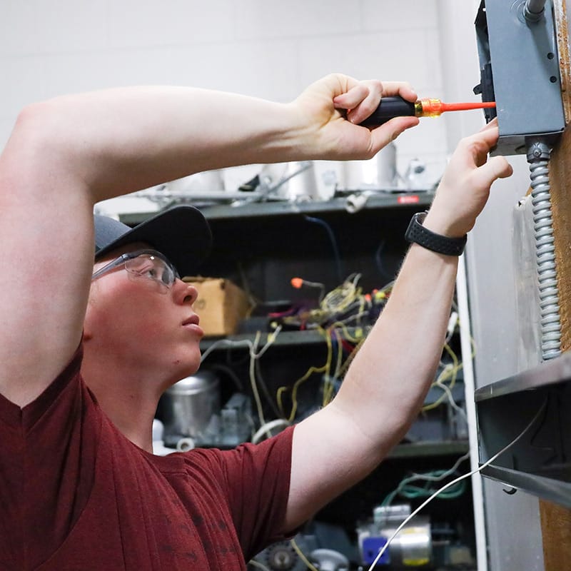 Student in safety glasses and a red shirt using a screwdriver on an electrical panel, with wires and equipment visible in the background.