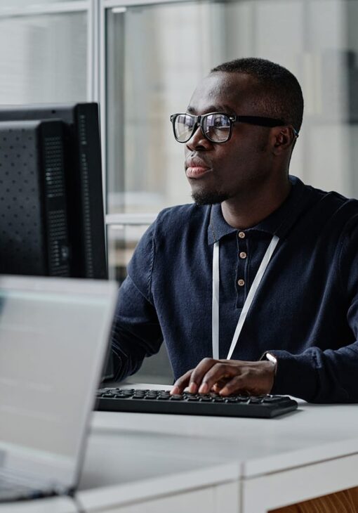 Man in glasses working intently at a desktop computer in a modern office setting.