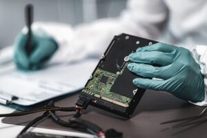 Gloved hands holding an open hard drive while taking notes on a clipboard, suggesting data recovery or forensic analysis.