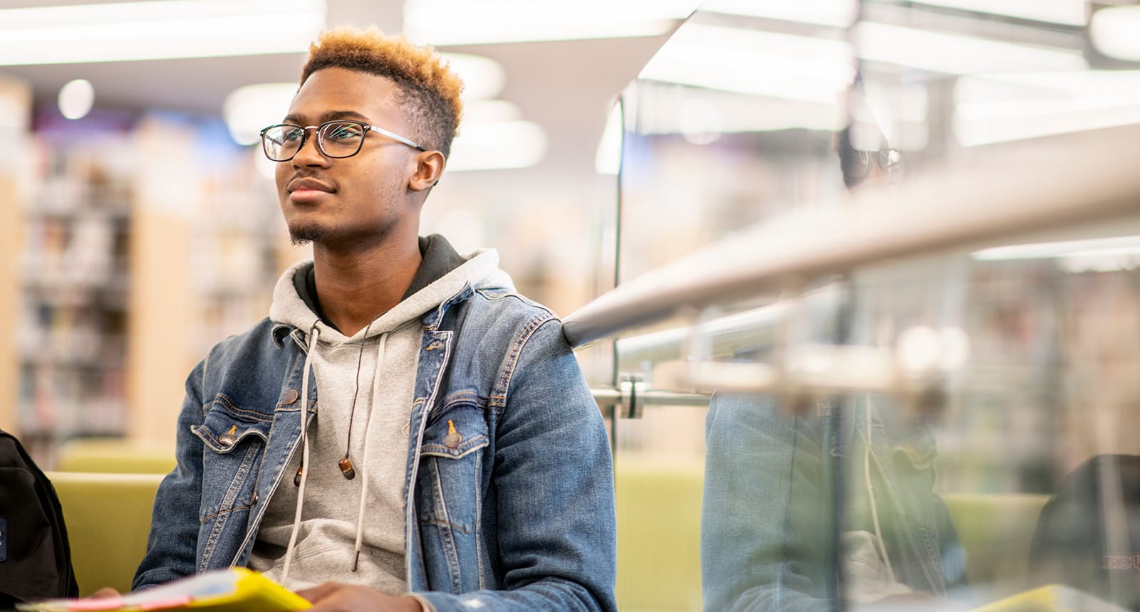 Student in glasses and a denim jacket sitting in a modern library holding a notebook
