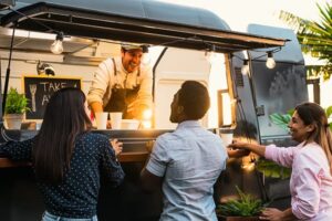 Smiling man inside a food truck serving drinks to three casually dressed customers standing outside at sunset.