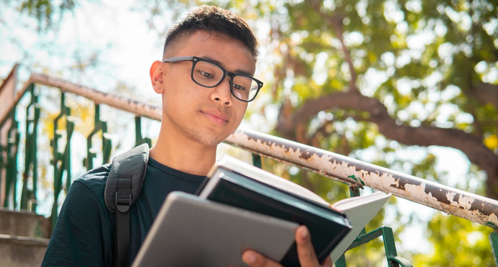 Student wearing glasses and a backpack reading a book outside on a sunny day.