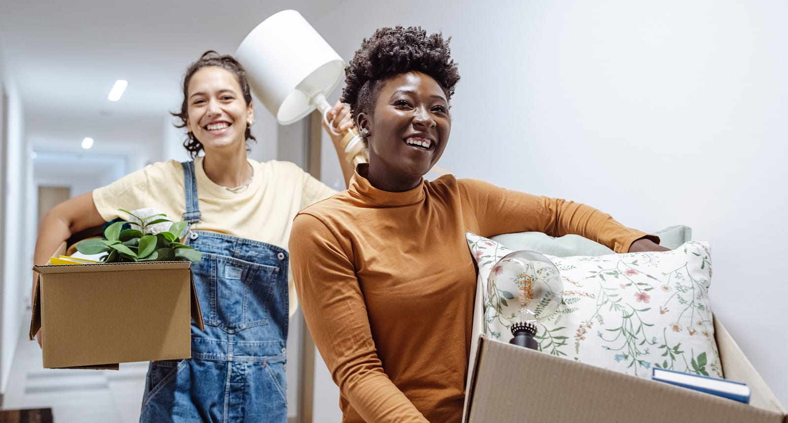 Two smiling young women carrying boxes and household items while moving into a dorm hallway.
