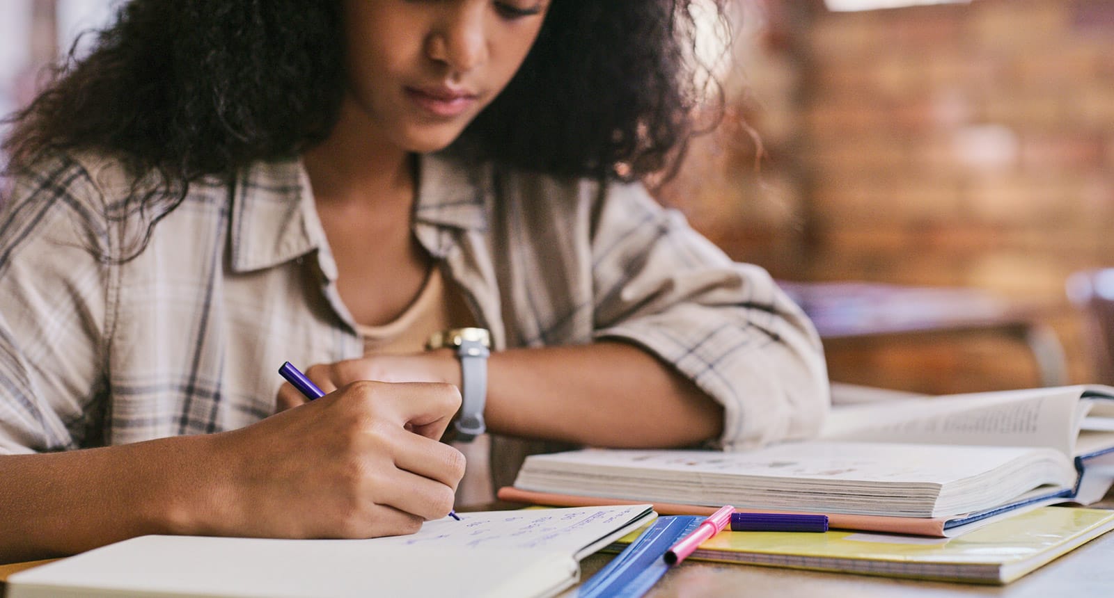 Student writing in a notebook with textbooks and colorful pens on a desk.