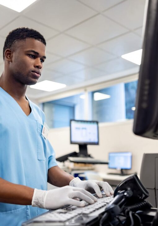 Medical professional in light blue scrubs and white gloves working at a computer station in a clinical setting, with multiple monitors and bright natural lighting.
