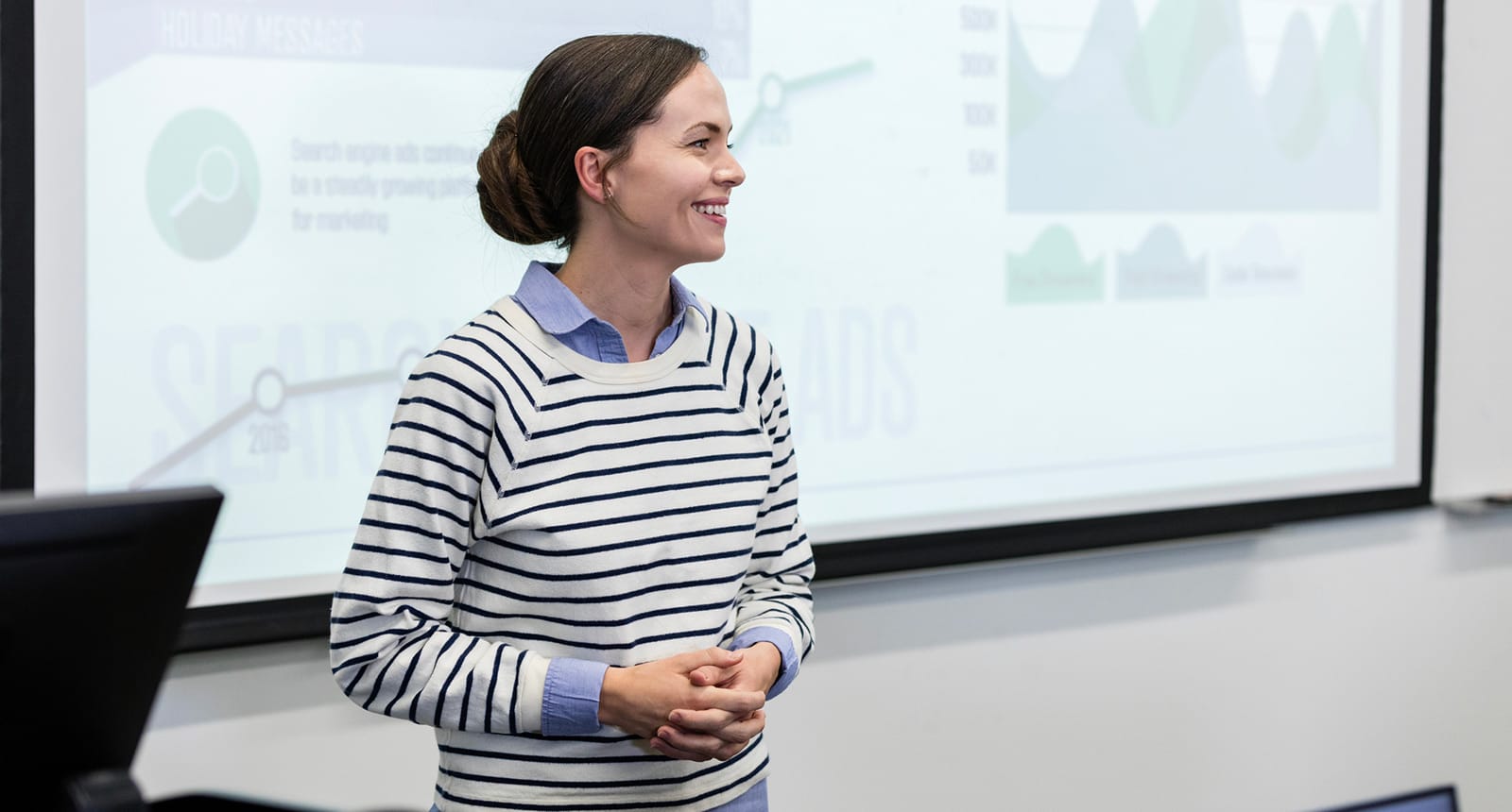 Smiling woman in a striped sweater stands in front of a projected presentation with charts and graphs.