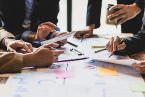 Hands of multiple people working with tablets, charts, and sticky notes during a business meeting at a table.