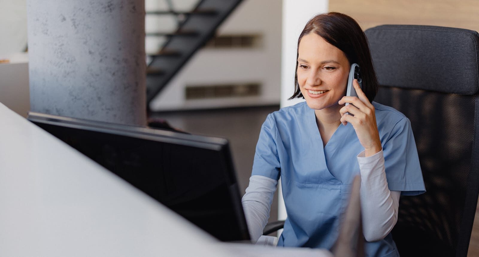 Smiling healthcare worker in light blue scrubs answering a phone call at a modern reception desk, with a computer monitor in front and a staircase in the background.